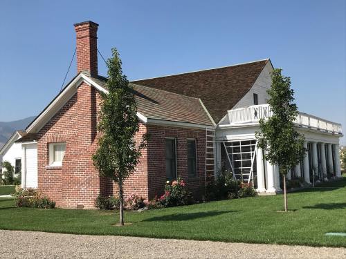 A brick house with a sloped roof, large windows, and a porch, surrounded by green grass and trees under a clear blue sky.