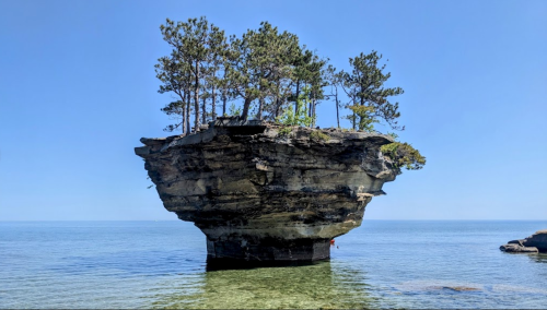 A small island with trees atop a large rock formation, surrounded by calm blue water under a clear sky.