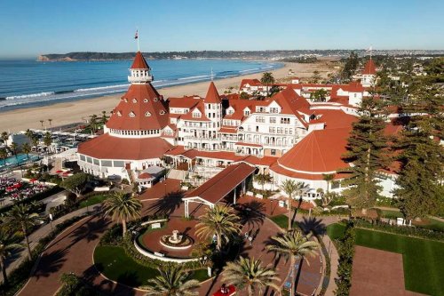 Aerial view of a historic hotel with red roofs, palm trees, and a beach in the background. Clear blue sky above.