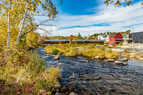 A scenic view of a river with a covered bridge, surrounded by autumn foliage and colorful buildings in the background.