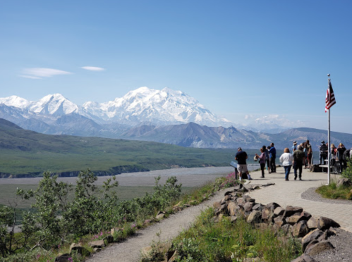 A scenic view of snow-capped mountains with visitors enjoying the landscape and an American flag in the foreground.