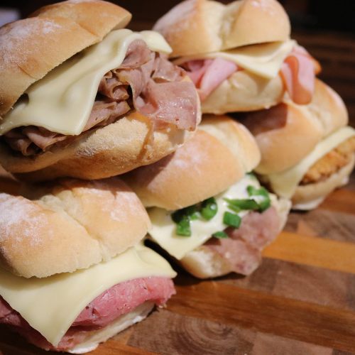 A stack of assorted sandwiches with various meats and cheese on soft rolls, arranged on a wooden surface.
