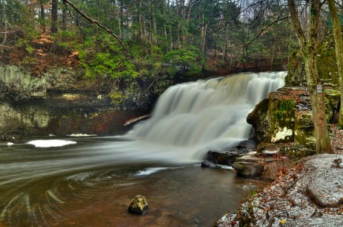 A serene waterfall cascades over rocks, surrounded by lush greenery and trees, creating a tranquil natural scene.