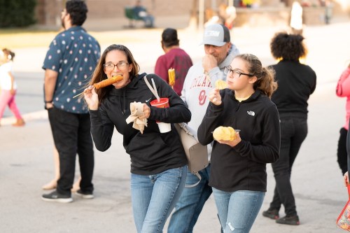 A woman and a girl enjoy snacks at an outdoor event, with people mingling in the background.