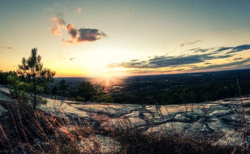 A panoramic view of a sunset over a landscape, with rocky terrain and trees silhouetted against the colorful sky.