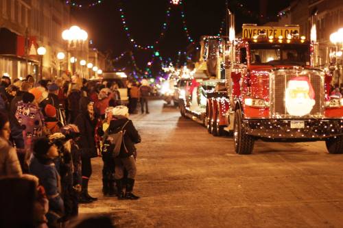 A festive night scene with a crowd watching a decorated truck with lights and a Santa face during a holiday parade.