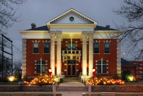 Historic brick building with columns, illuminated by festive lights and decorations, set against a twilight sky.