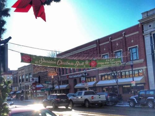 A festive banner for the Sheridan Christmas Stroll hangs over a downtown street, with shops and cars visible below.