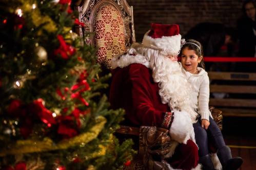 A young girl sits next to Santa Claus, sharing a joyful moment by a decorated Christmas tree.