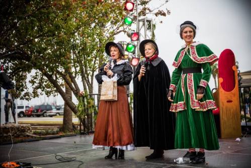 Three women in historical costumes stand on stage, holding microphones, with festive lights and trees in the background.
