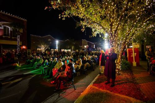 A crowd gathers under festive lights at night, enjoying an outdoor event in a charming town setting.