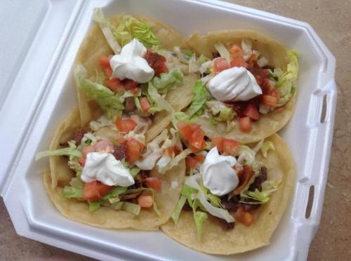 Four tacos in a takeout container, filled with lettuce, tomatoes, beef, and topped with sour cream and salsa.