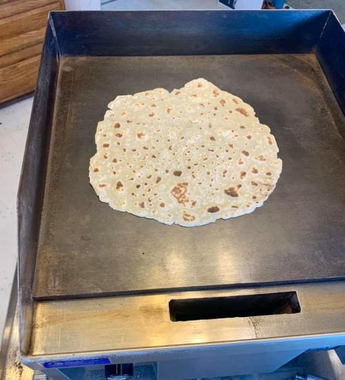 A large, round flatbread cooking on a hot griddle, showing a golden-brown surface with some charred spots.