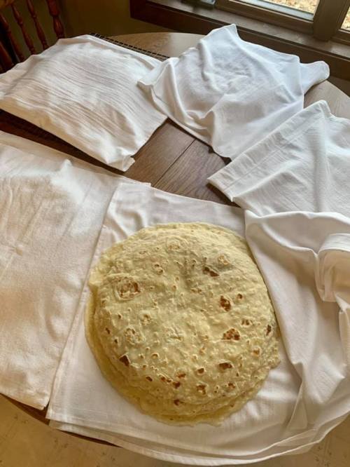 A large flatbread resting on a white cloth, surrounded by more cloths on a wooden table. Natural light from a window.