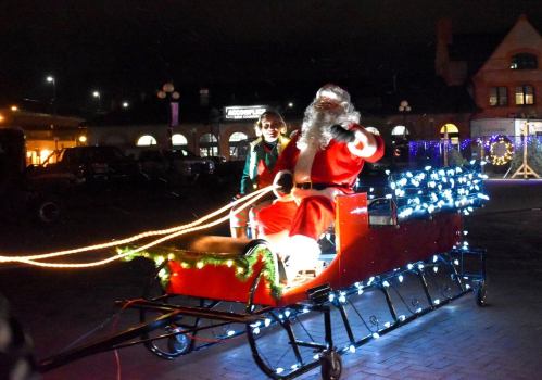Santa Claus in a decorated sleigh, smiling and waving, with festive lights, in a nighttime holiday setting.