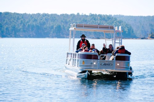 A group of people on a pontoon boat waves while cruising on a sunny lake surrounded by trees.