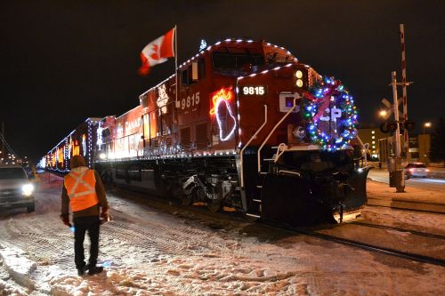 A brightly lit train decorated for the holidays, with a person in an orange vest walking nearby in a snowy setting.