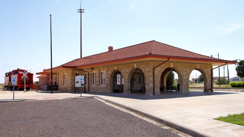 Historic stone train station with a red roof, featuring arched entrances and a vintage train car nearby.