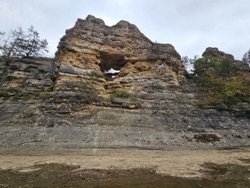 A rocky cliff with a large natural arch, surrounded by trees and a cloudy sky.
