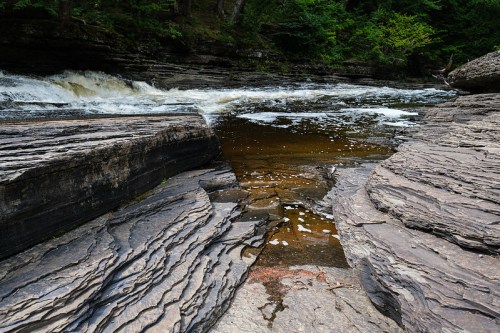 A rocky riverbank with layered stones and flowing water, surrounded by lush green trees.