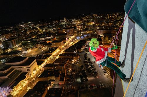 A person dressed as the Grinch rappels down a building at night, overlooking a city decorated with holiday lights.