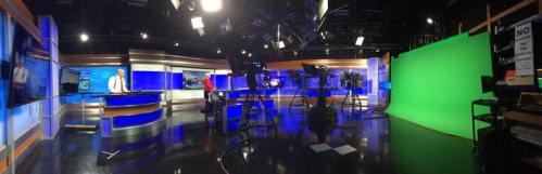 A panoramic view of a TV news studio featuring a news desk, cameras, and a green screen backdrop.