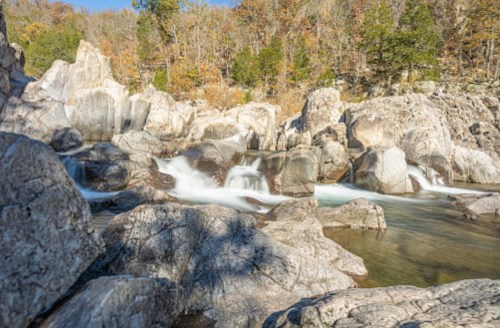 A serene landscape featuring a flowing river over rocky terrain, surrounded by autumn foliage and trees.