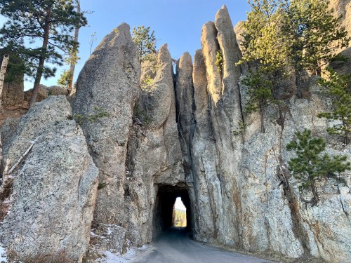A narrow road passes through a rocky tunnel surrounded by tall, jagged rock formations and pine trees.