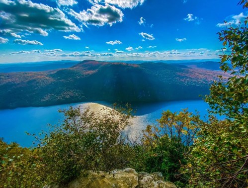 Scenic view of a lake surrounded by colorful autumn foliage and mountains under a bright blue sky with fluffy clouds.