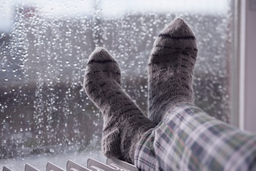 Cozy feet in striped socks resting on a radiator, with raindrops on a window in the background.