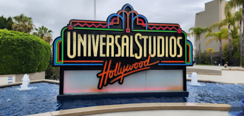 Colorful Universal Studios Hollywood sign in front of a water feature, surrounded by palm trees and a cloudy sky.