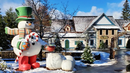 A large snowman statue with a green hat and scarf stands outside a snowy building, surrounded by snow-covered trees.