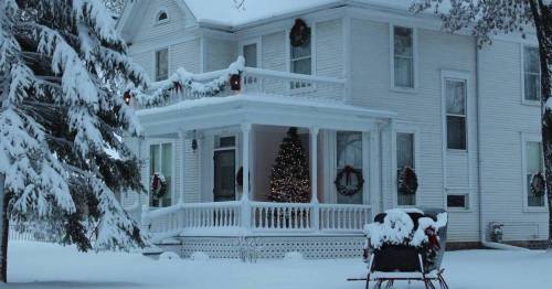 A snowy white house decorated for the holidays, featuring a Christmas tree visible through the front window.