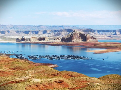 A scenic view of a lake surrounded by red rock formations and distant cliffs under a clear blue sky.
