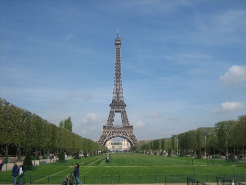 Eiffel Tower towering over a green park under a blue sky with scattered clouds. People stroll along the pathways.