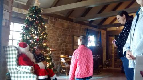 A cozy room decorated for Christmas, with a child talking to Santa by a tree, while another child and an adult watch.