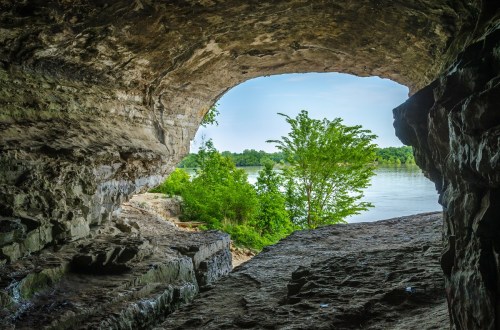 View from a cave opening overlooking a river and lush greenery on the opposite bank.