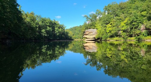 A serene river scene with lush green trees and a large rock formation reflected in the calm water under a clear blue sky.
