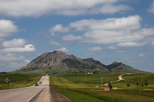 A scenic view of a highway leading towards a large, rocky mountain under a partly cloudy sky. Green fields surround the road.