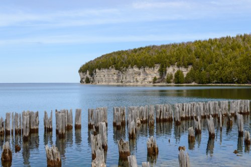 A serene lake scene with weathered wooden posts in the foreground and a lush, rocky cliff in the background.