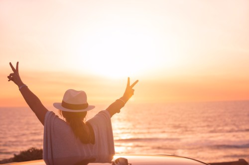 Silhouette of a person in a hat, making peace signs against a sunset over the ocean.