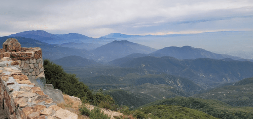 A panoramic view of mountains and valleys under a cloudy sky, with a stone wall in the foreground.