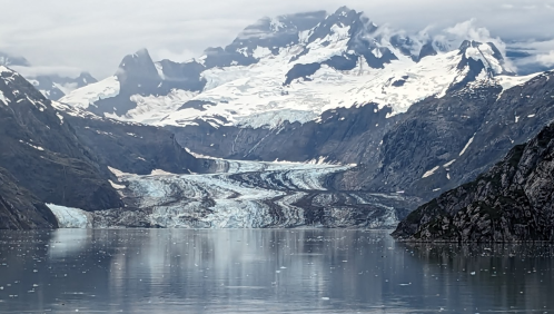 A stunning glacier flows down a mountain valley, surrounded by snow-capped peaks and calm waters reflecting the landscape.