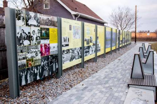 A long display of historical photos and information panels along a pathway, with benches and a house in the background.