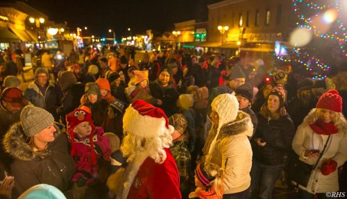 A festive crowd gathers at night, with people in winter attire and a Santa figure among them, celebrating the holiday season.