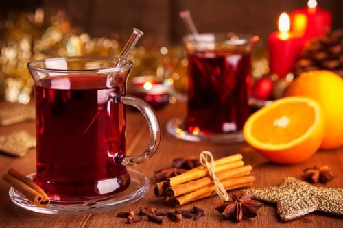 Warm spiced drinks in glass mugs, garnished with cinnamon sticks and orange slices, set against a festive background.