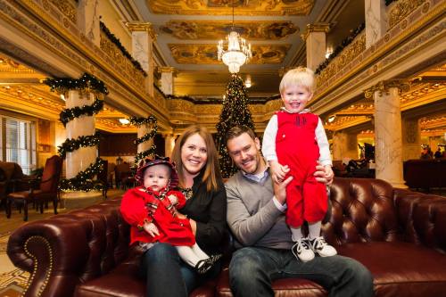 A family of four poses on a couch in a festive, elegantly decorated lobby with a Christmas tree in the background.