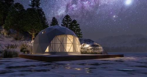 A geodesic dome cabin by a frozen lake, illuminated under a starry night sky with trees in the background.