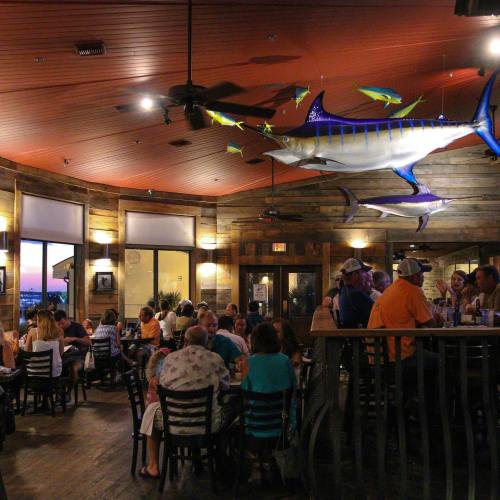 A lively restaurant interior with wooden decor, large fish sculptures, and diners enjoying their meals at sunset.