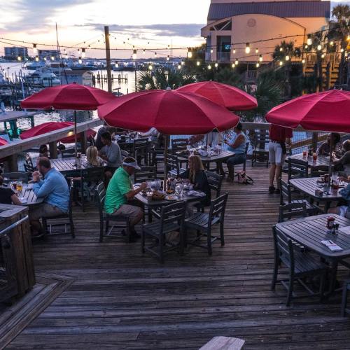 Outdoor dining area with red umbrellas, people enjoying meals, and a waterfront view at sunset.